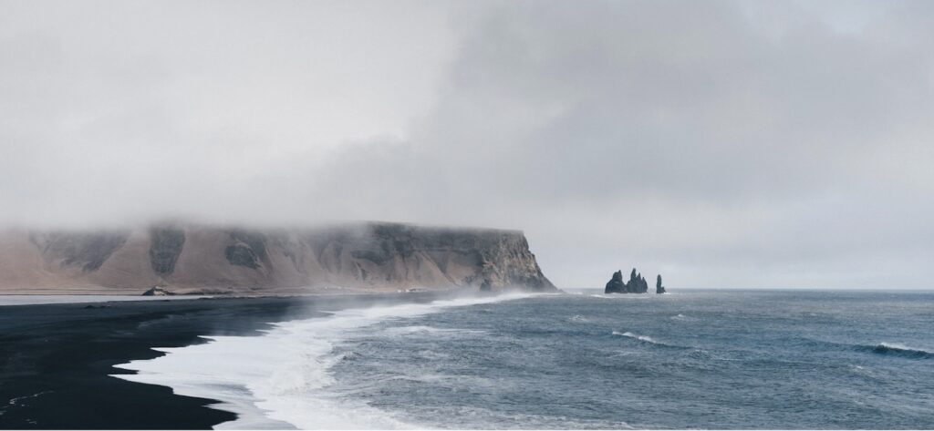 A misty coastal scene featuring a black sand beach, rolling waves, a cliff, and sea stacks in the distance beneath a cloudy sky.