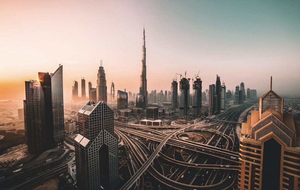 Aerial view of a modern cityscape with tall skyscrapers and intricate highway interchanges at sunset.