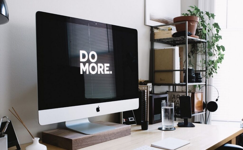 A desktop computer with "DO MORE." displayed on the screen sits on a tidy office desk with a shelving unit, plants, and various office items in the background.