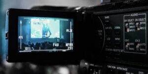 **Alt Text:** The image shows the display screen of a video camera recording a person speaking on stage to a seated audience.