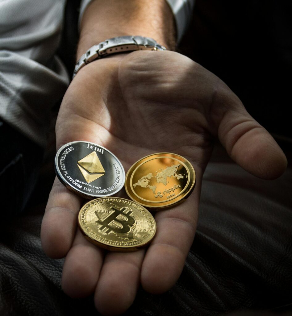 **Alt Text:** A hand holding three cryptocurrency coins: Ethereum, Ripple, and Bitcoin.