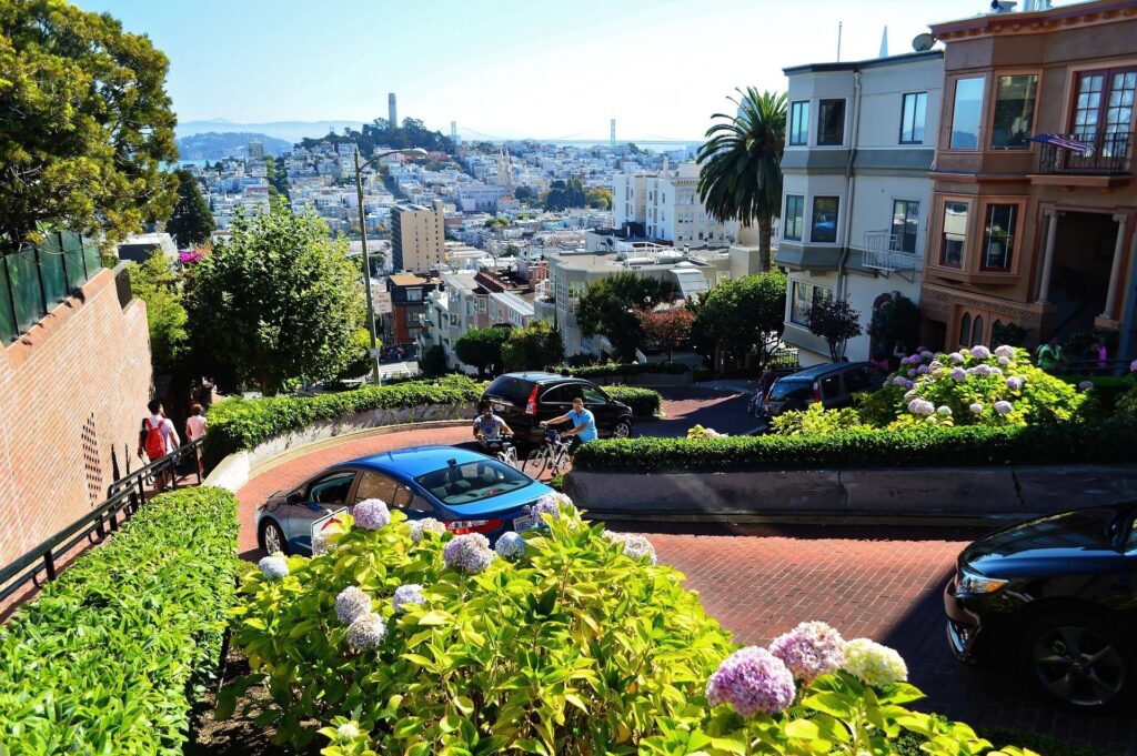 Cars and cyclists navigate the winding Lombard Street in San Francisco, surrounded by lush greenery and overlooking the cityscape.