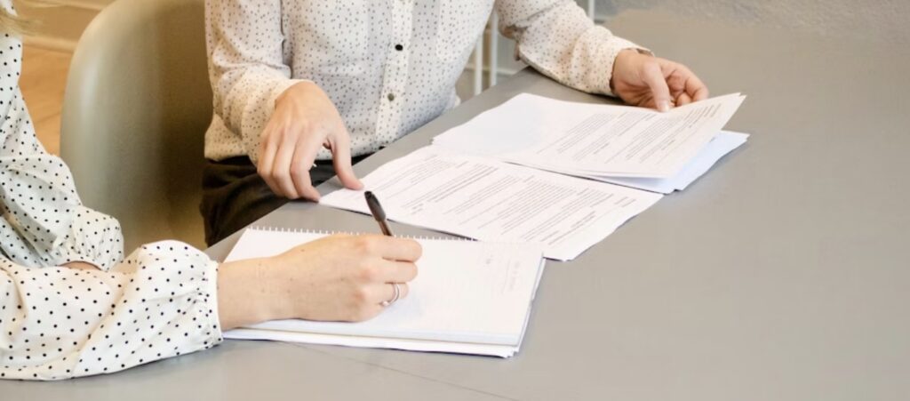 **Alt text:** Two people sitting at a table reviewing documents, with one person writing notes in a notebook.