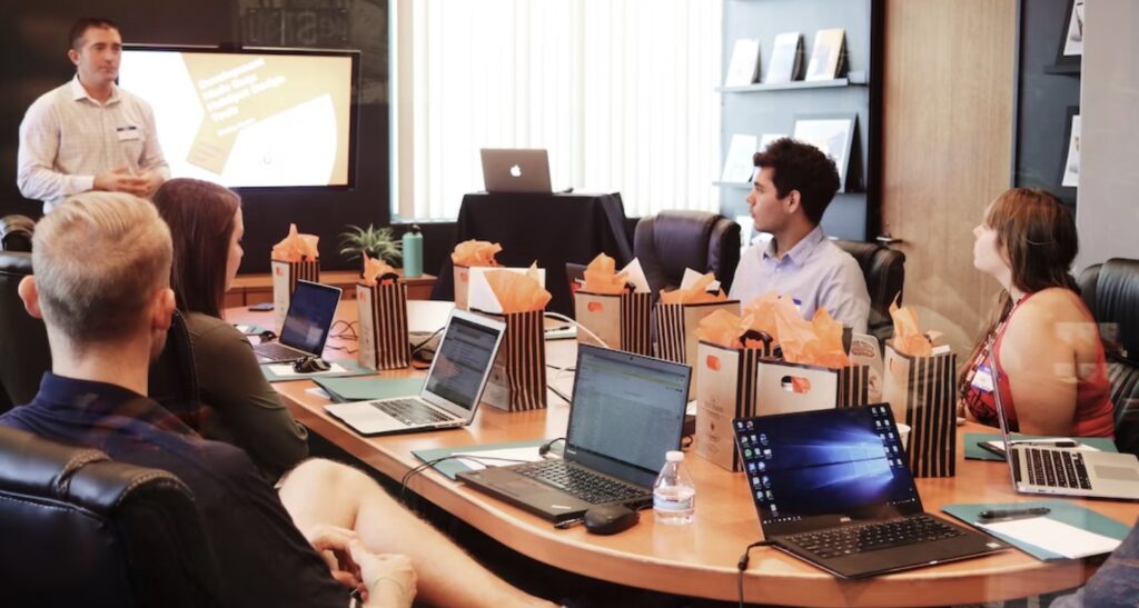 A group of people in a conference room are listening to a presentation, with laptops and gift bags on the table.