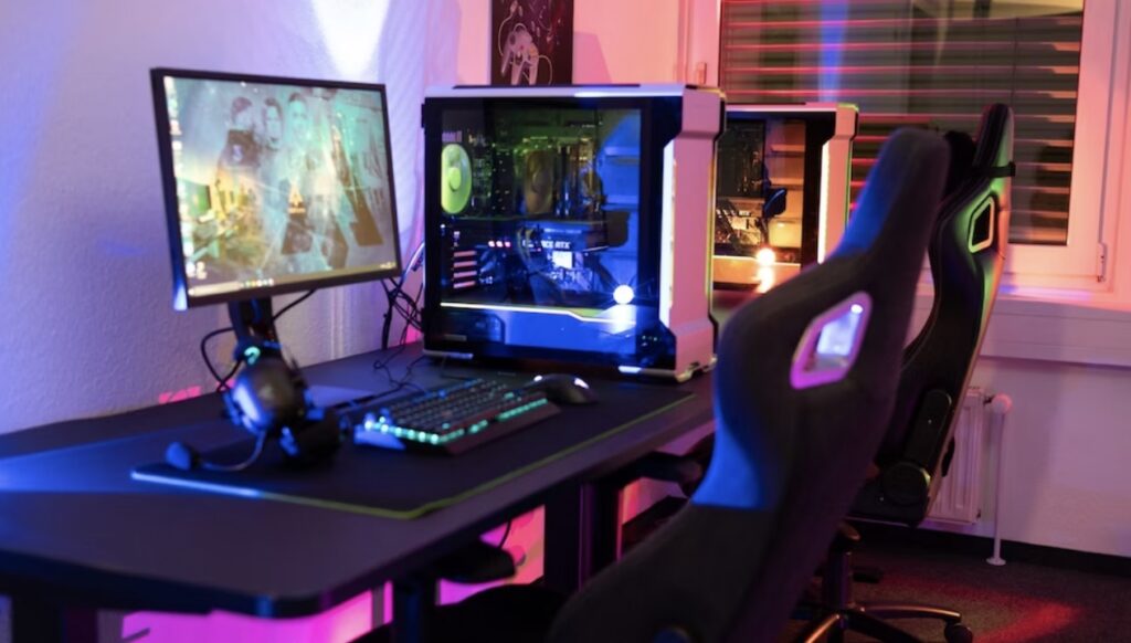 A gaming setup featuring a dual-monitor computer, a backlit keyboard, a gaming chair, and a desktop tower with glowing internal components.