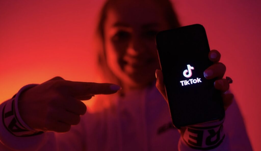 A person points to a smartphone displaying the TikTok logo against a red-lit background.
