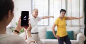 **Alt text:** A person holds up a phone displaying the TikTok logo while two older adults dance in the background.