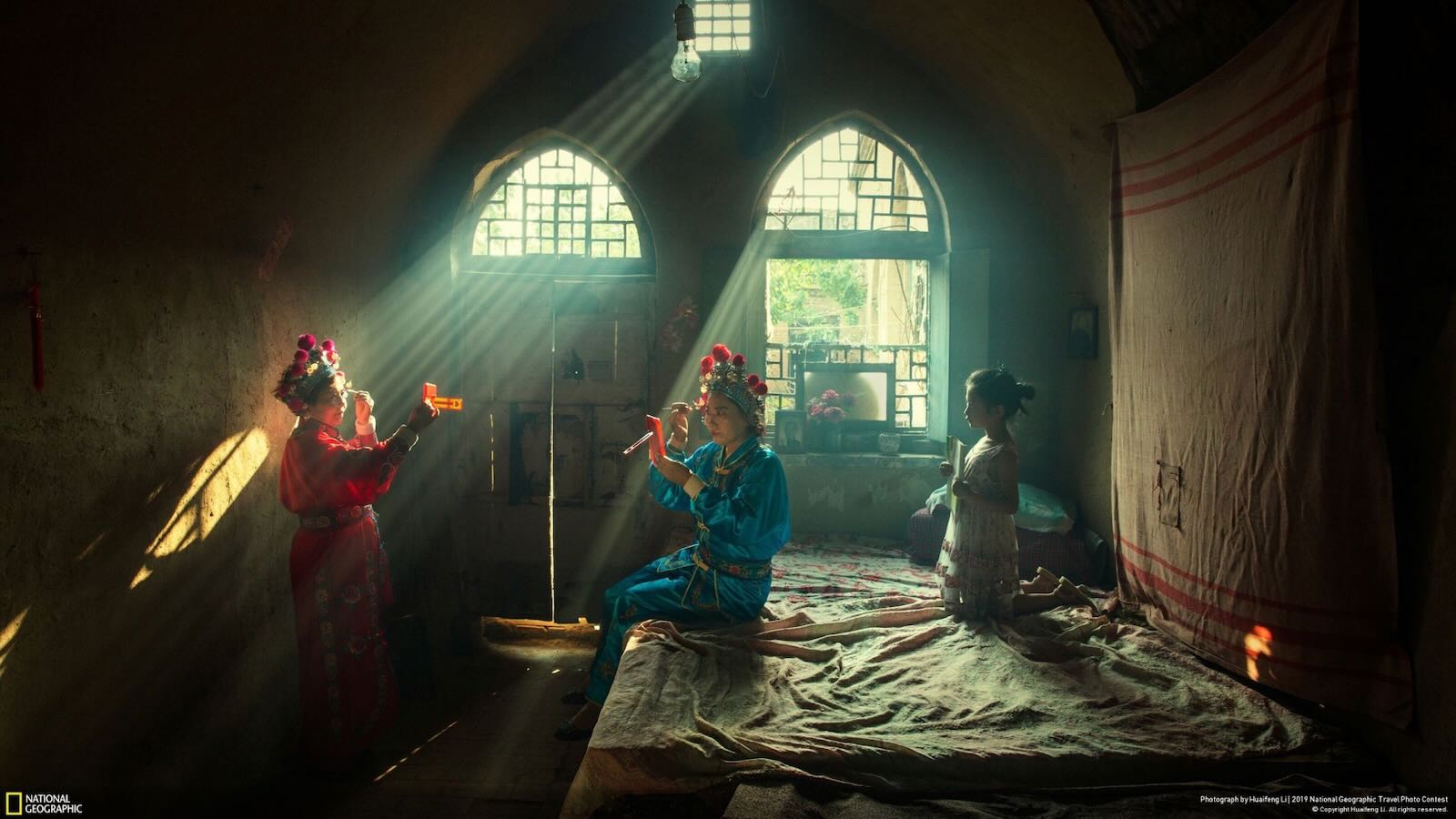 Two people dressed in traditional costumes apply makeup in a dimly lit room with sunlight streaming through arched windows, observed by a child.