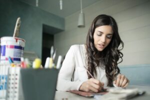 A woman with long hair is sitting at a table, focused on writing or drawing with a pen, surrounded by art supplies.