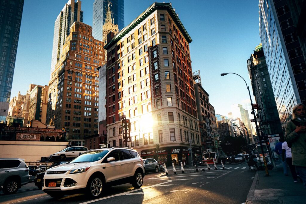 A busy city street with cars and pedestrians, surrounded by tall buildings reflecting the sunlight.