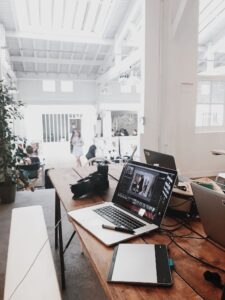 A wooden table with a laptop displaying photo editing software, a digital drawing tablet, and a camera, situated in a bright, spacious room with high ceilings and visible people in the background.