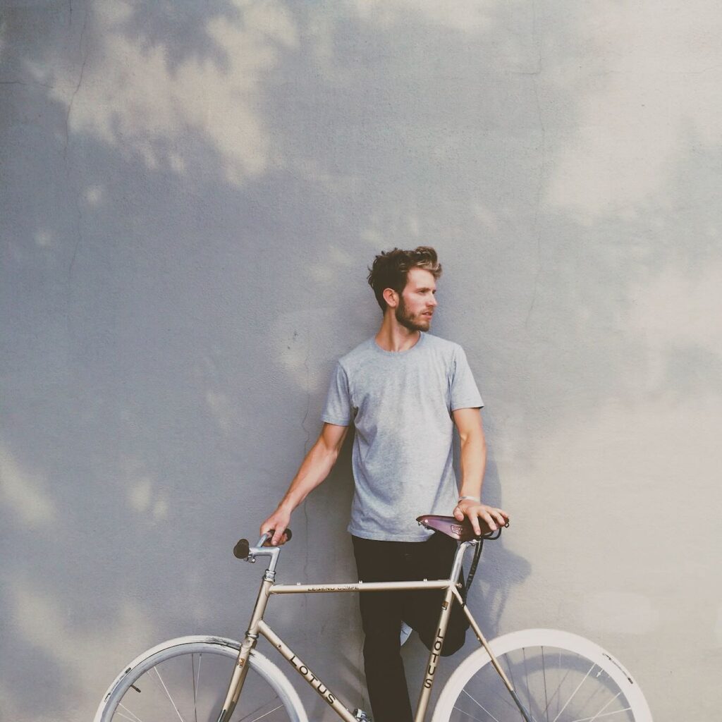 **Alt text:** A man in a gray t-shirt leans against a wall with a bicycle, looking to the side.
