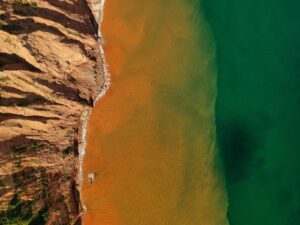 **Alt Text:** Aerial view of a coastal meeting point where red cliff formations meet orange-tinged shallow water transitioning into deep green ocean.