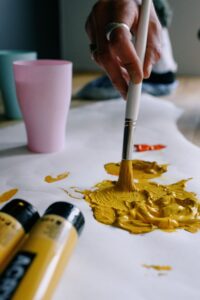 A person uses a paintbrush to mix yellow paint on a surface, with paint tubes and cups nearby.