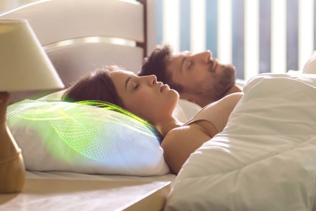 A man and woman are lying in bed asleep with a glowing green and blue wave pattern on the woman's pillow.