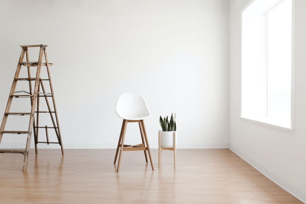 **Alt Text:** A minimalist room with a wooden ladder, a white chair, and a small potted plant near a window.