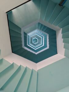 **Alt Text:** An upward view of a spiral staircase with alternating turquoise and white steps forming a geometric pattern.