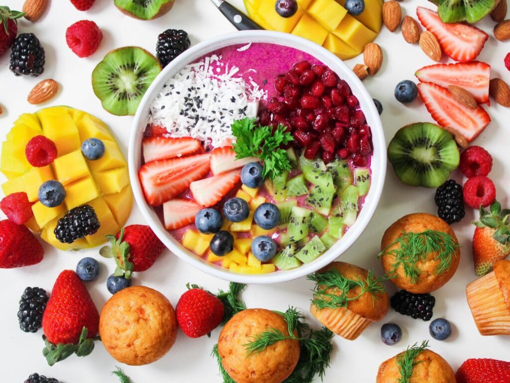 A colorful smoothie bowl topped with a variety of fresh fruits, surrounded by scattered fruit pieces, almonds, and small muffins.