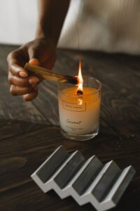 **Alt text:** A hand lighting a coconut-scented candle with a burning stick on a wooden table.