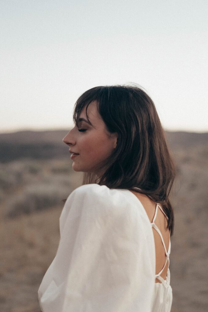 A woman in a white dress stands outdoors with her eyes closed, against a blurred natural background.
