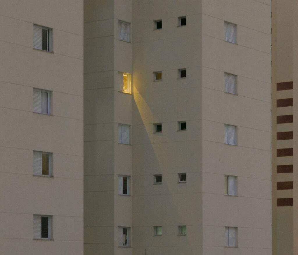 A tall apartment building with one illuminated window casting light onto another window, while the rest remain dark.