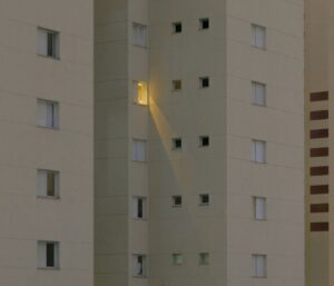 A tall apartment building with one illuminated window casting light onto another window, while the rest remain dark.
