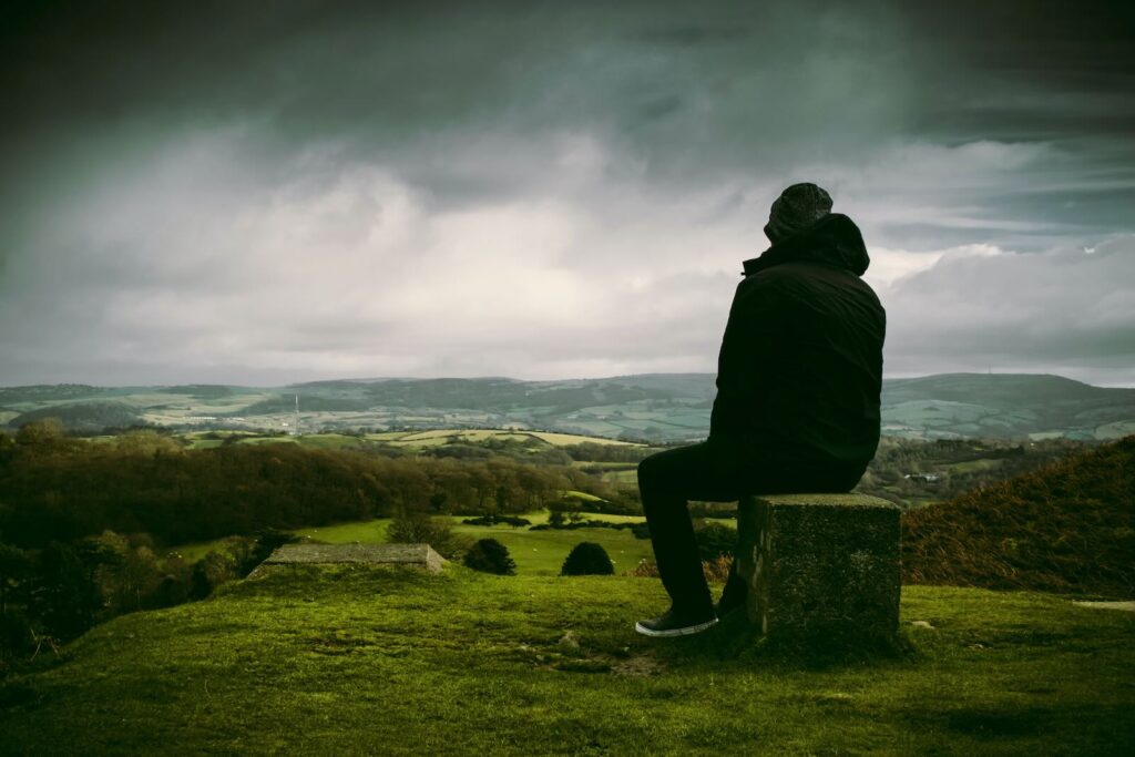 **Alt text:** A person in a jacket sits on a stone block, overlooking a vast, green landscape under a cloudy sky.