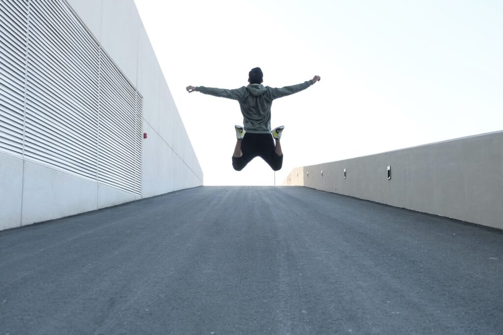 **Alt text:** A person in a hoodie jumps with arms outstretched on an ascending asphalt pathway between two walls.