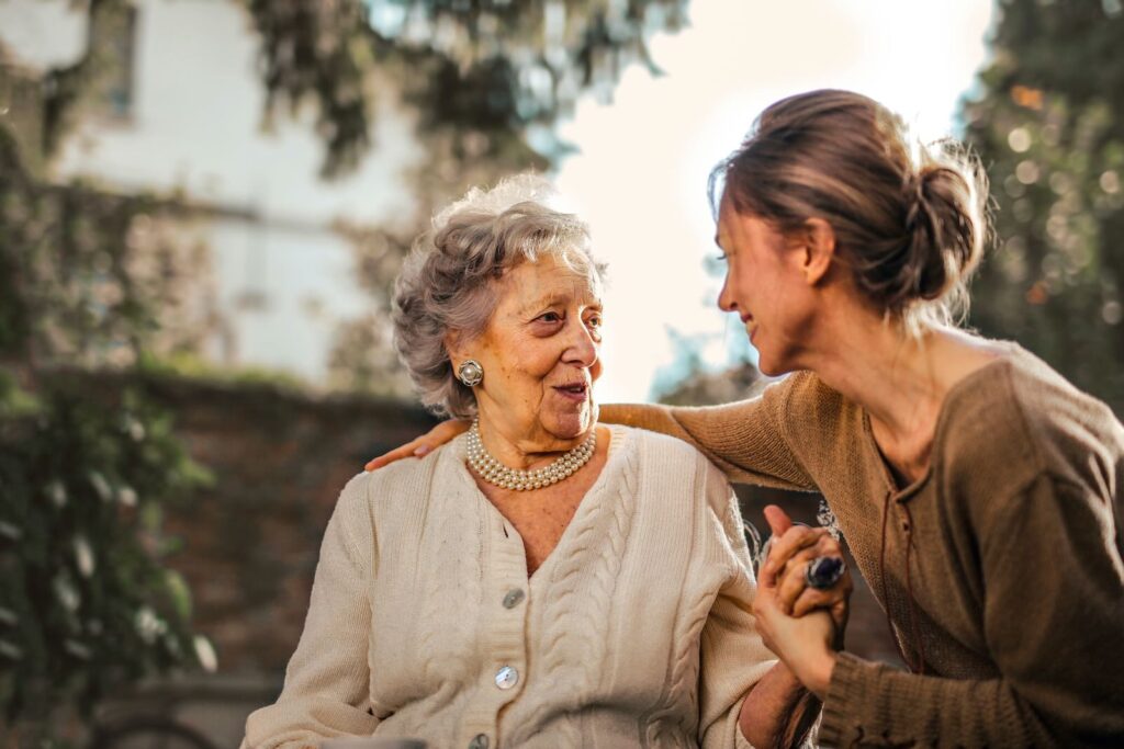 A smiling elderly woman in a beige sweater is lovingly embraced by a younger woman in an outdoor setting.