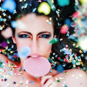 **Alt text:** A woman with dramatic eye makeup holds a pink lollipop amidst colorful confetti scattered around her.