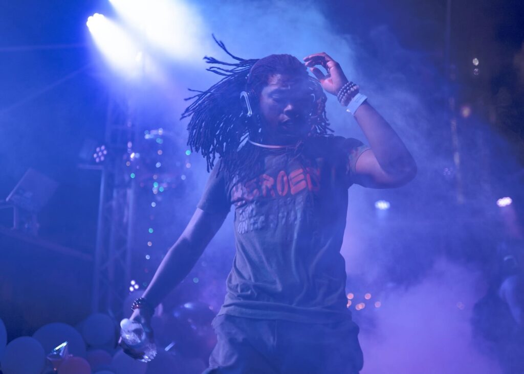 **Alt Text:** A person with long dreadlocks dances energetically on stage under vibrant purple and blue lighting, holding a water bottle.