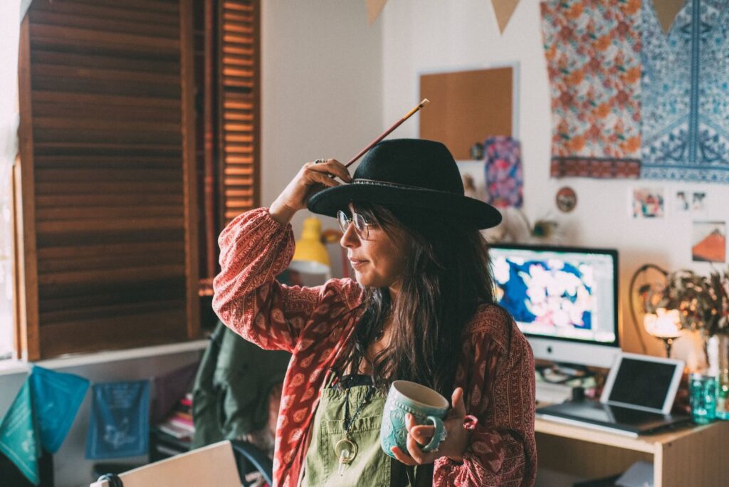 **Alt Text:** A woman wearing a hat and glasses holds a pencil in one hand and a mug in the other, standing in a colorful room with a computer in the background.