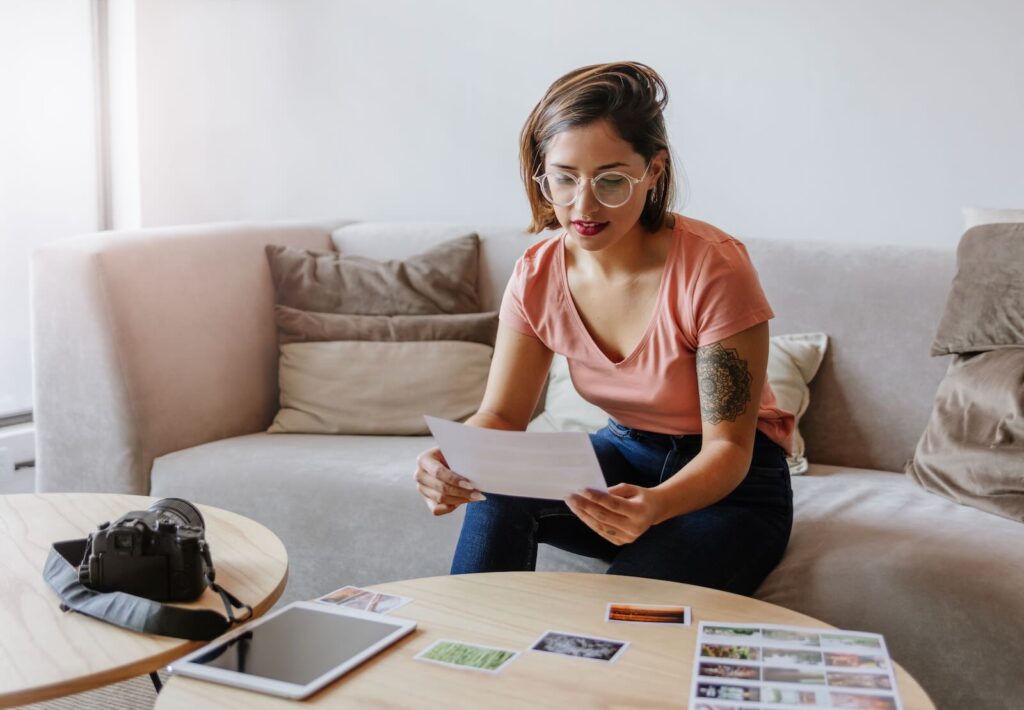 **Alt text:** A woman sitting on a sofa looks at a paper in her hand, with a camera, tablet, and photos on the table in front of her.