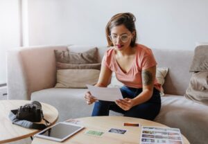 **Alt text:** A woman sitting on a sofa looks at a paper in her hand, with a camera, tablet, and photos on the table in front of her.