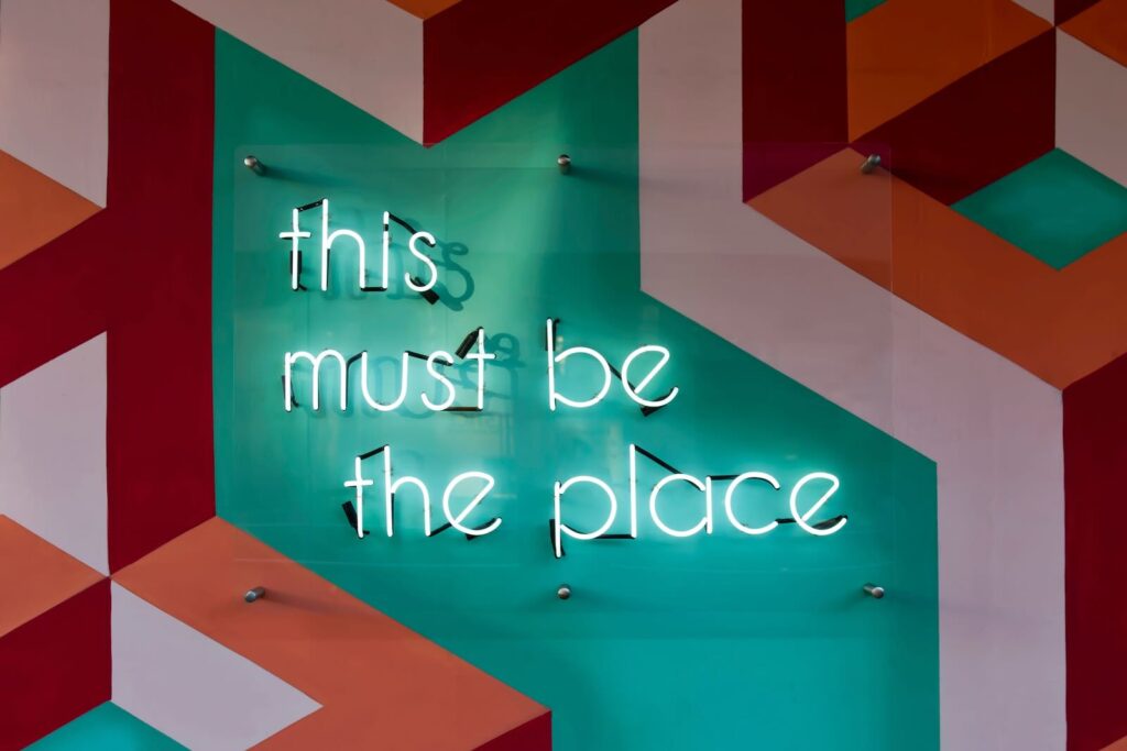 **Alt text:** A neon sign reading "this must be the place" is mounted on a colorful geometric-patterned wall.