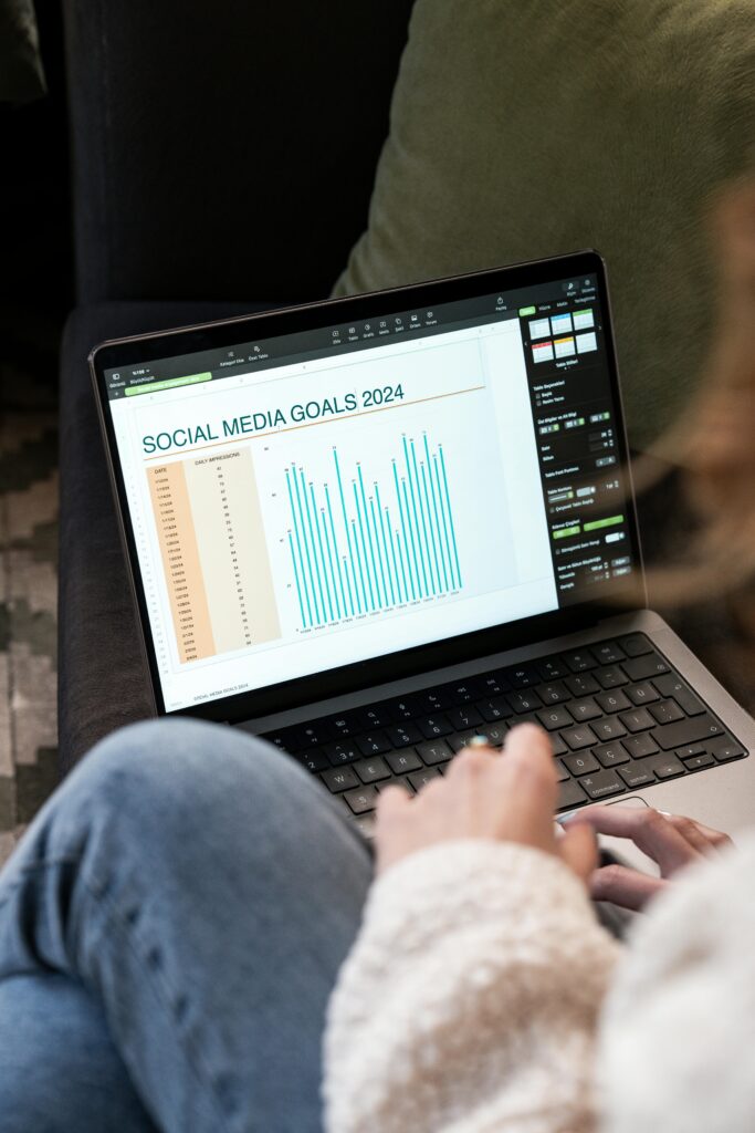 A person sits on a couch using a laptop displaying a spreadsheet titled "Social Media Goals 2024" with a bar chart.
