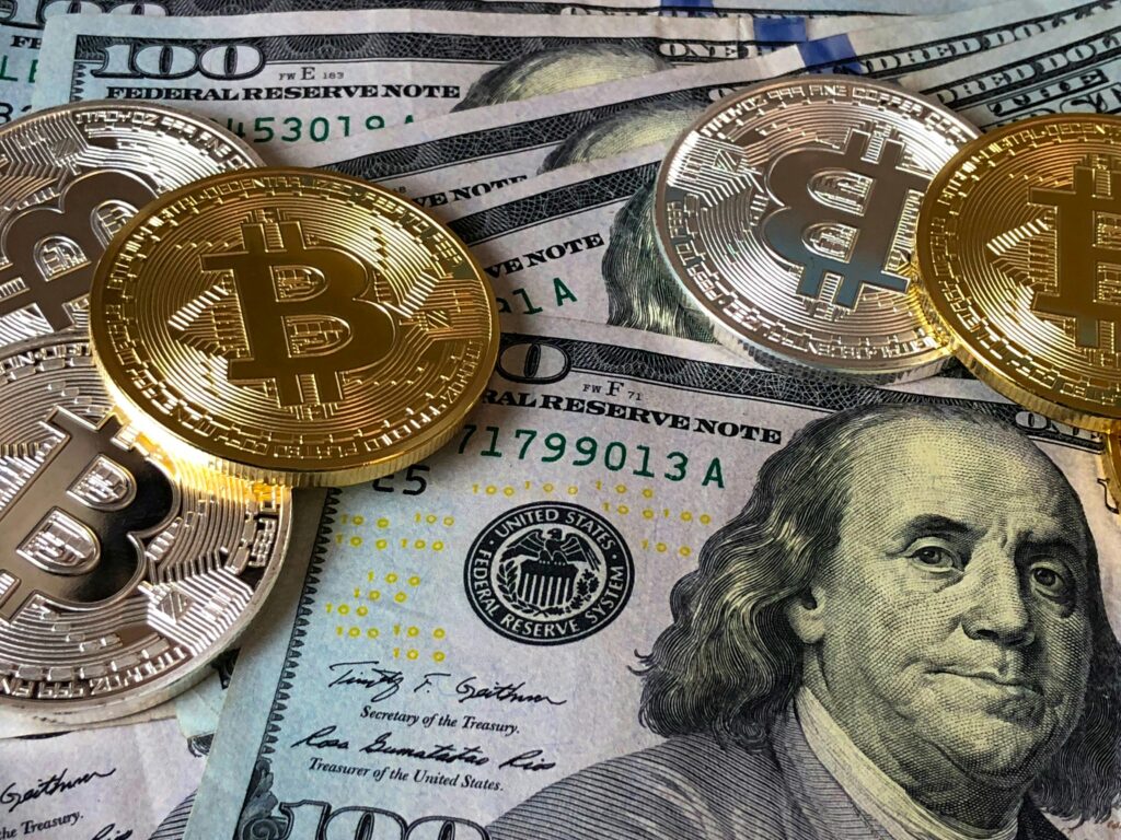 **Alt text:** Gold and silver Bitcoin coins are laid on top of several one hundred dollar bills.