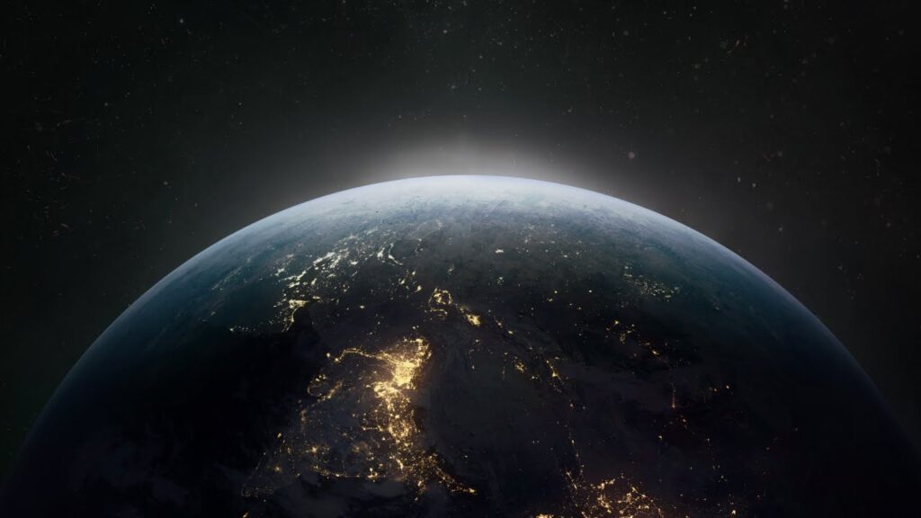 **Alt text:** A view of Earth from space showing illuminated city lights on the dark side as the sun begins to rise over the horizon.