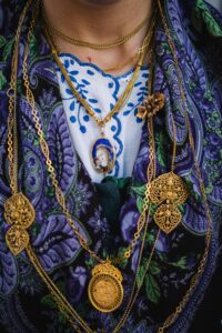 **Alt Text:** A person wearing a traditional outfit adorned with multiple gold necklaces, including intricate filigree designs and a pendant featuring an image of a saint or religious figure.