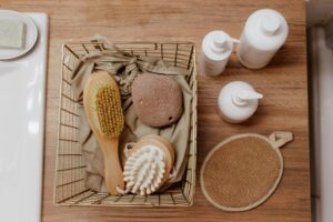 **Alt text:** A bathroom essentials set on a wooden countertop includes a wire basket containing a brush, pumice stone, and massager, alongside three white pump bottles and an exfoliating mitt.