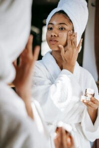 A person wearing a white bathrobe and a towel on their head applies face cream while looking in the mirror.