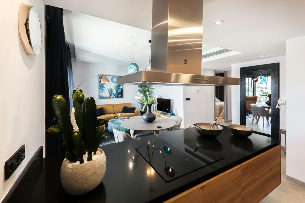 A modern kitchen with a black countertop, stainless steel range hood, and a cactus plant in a white pot, overlooking an open-plan living room with a yellow sofa.