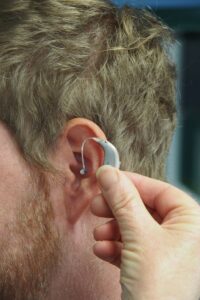 **Alt Text:** A hand is fitting a hearing aid behind a man's ear.