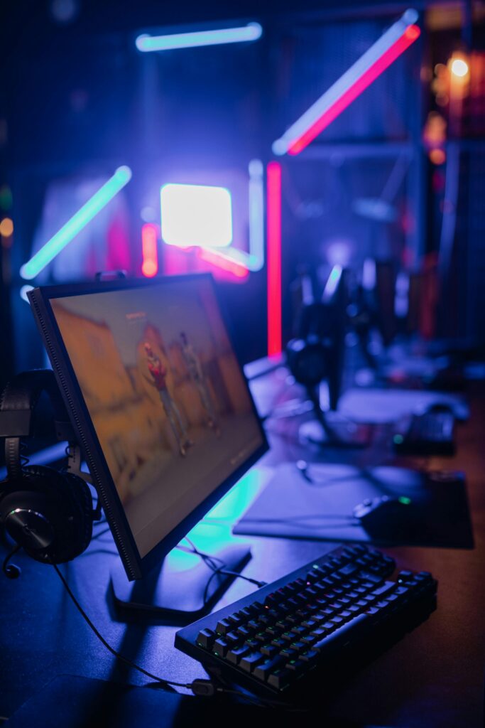 A gaming setup with a monitor, keyboard, and headphones, illuminated by neon blue and red lights in a dimly lit room.
