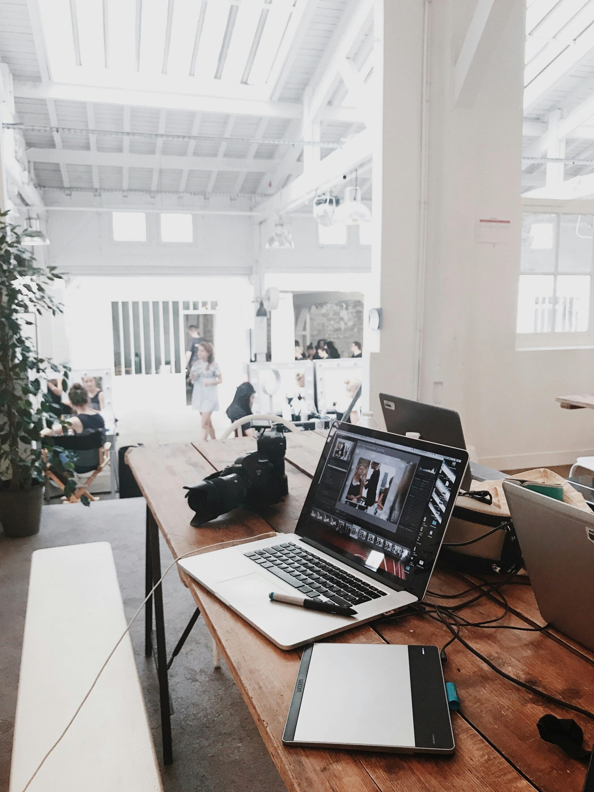A bright, modern workspace with a wooden table holding a laptop, camera, and tablet, in an open room with high ceilings and people in the background.