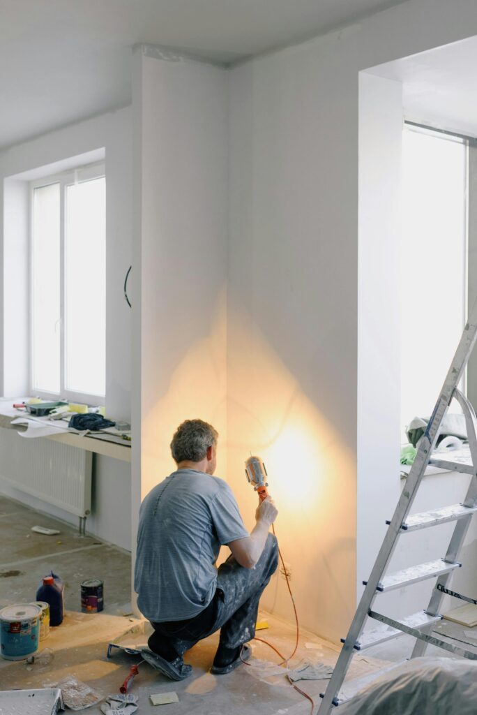 A person uses a hand tool to work on a wall in a partially renovated room with scattered tools and a stepladder.