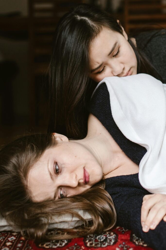 A woman lying on a carpet, wrapped in a blanket, with another woman resting beside her in a comforting manner.