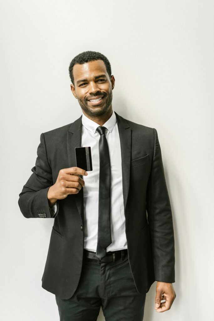 A man in a black suit and tie smiles while holding a credit card.