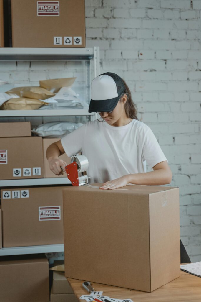 A person wearing a cap is sealing a large cardboard box with tape in a warehouse filled with other boxes.