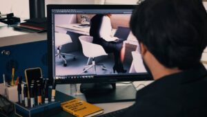 A person is sitting at a desk, looking at a computer screen displaying an image of a person working on a laptop in an office setting.
